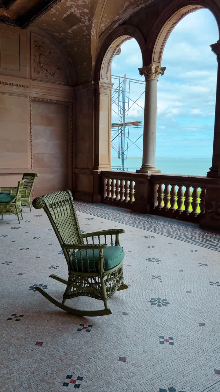 Innenbalkon mit Bogenfenstern und Meerblick, mit Korbstuhl. Luxuriöse Loggia eines Anwesens mit Mosaikfliesenboden und grünem Korbschaukelstuhl mit Meerblick.