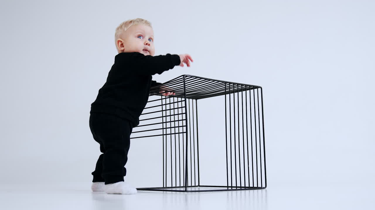 Blond baby in black suit and white socks stands near the metal wire cube. Kid taps his hand by the cube and smiles. Low angle view. White backdrop.