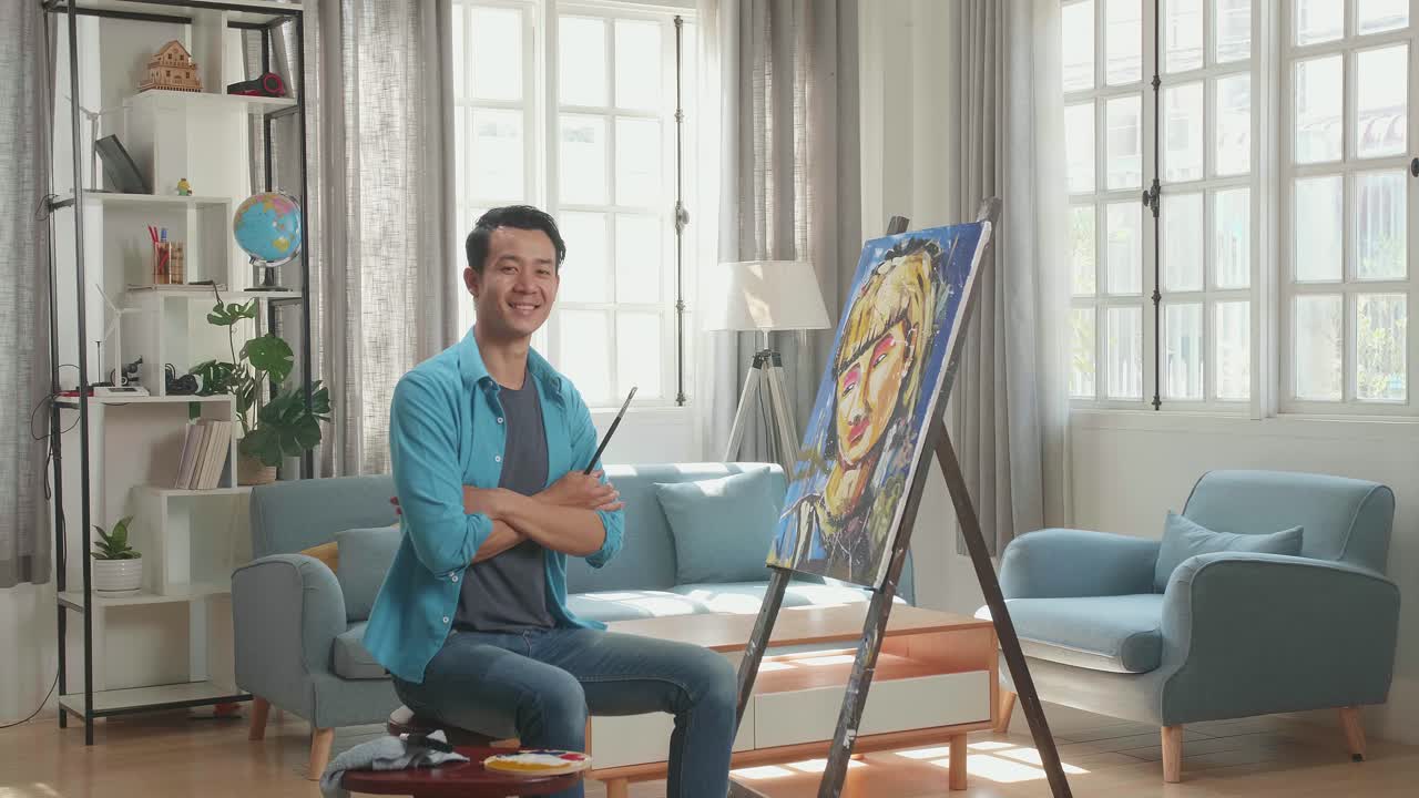 Asian Artist Man Sitting On Round Wooden Chair Without Backrest Holding Paintbrush Mixed Colour, Crossing His Arms, And Smiling After Finish Painting A Girl On The Canvas