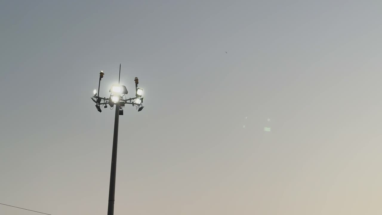 Lampu jalan tiang tinggi bersinar melawan langit malam, satu lampu berkedip-kedip karena kerusakan, menyoroti sistem penerangan jalan perkotaan dan masalah pemeliharaan saat ini.