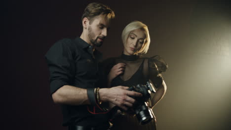 hombre fotógrafo mostrando fotos de la sesión de fotos a la joven modelo rubia en la pantalla de la cámara en el estudio oscuro