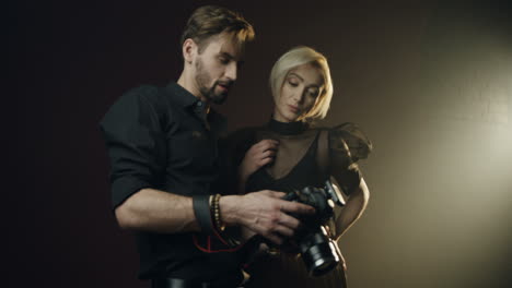 photographer man showing photos of photoshoot to the young blond woman model on the camera screen in the dark studio