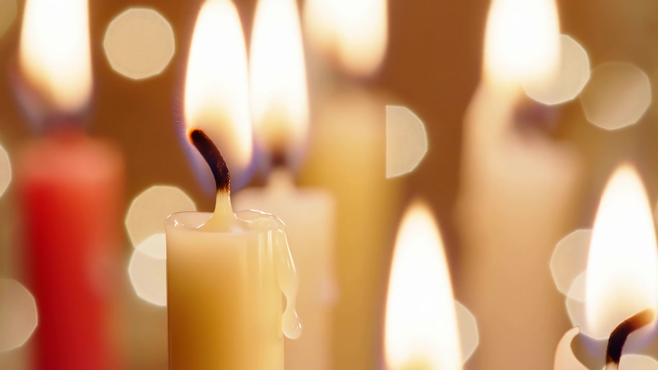 Candela color crema in primo piano con stoppino annerito e arricciato e una goccia di cera viscosa che pende dal bordo fuso, mentre le candele accese circostanti proiettano fiamme alte e sfocate, bokeh rotondo, bagliore morbido e luce calda.