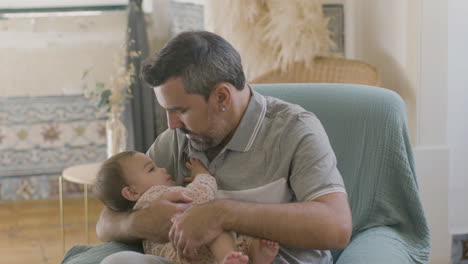 Caring-Dad-Sitting-In-Armchair-And-Cradling-His-Cute-Baby-Girl-At-Home