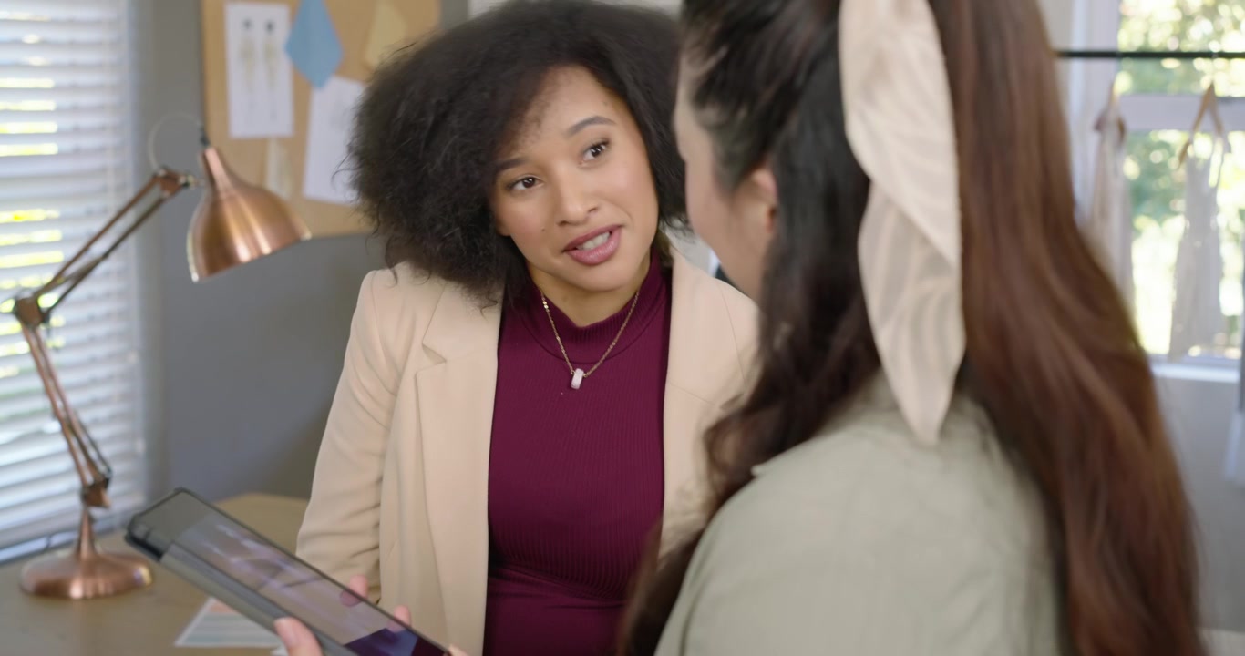 Diverse female coworkers presenting tablet, leaning, tapping and reviewing designs by copper lamp
