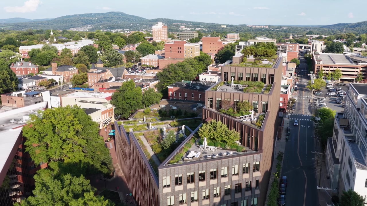 Zahradní terasy moderní budovy nad ulicemi centra Charlottesville ve Virginii, s parkovacími domy, cihlovými bloky a pohořím Blue Ridge v pozadí. Široký boční letecký záběr