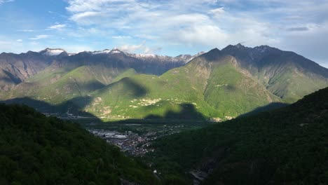 ariel ascending pedestal shot of shadow speckled town in the valley of giant magnificent mountains with snow covered mountains in the background and partly cloudy sky