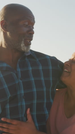 una pareja afroamericana mayor sonriente abrazándose y caminando por una playa soleada