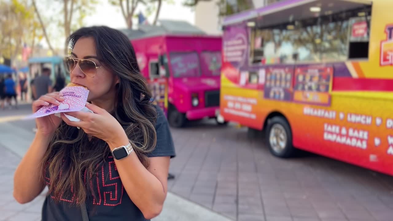mujer hispana en primer plano comiendo comida de un camión de comida - fondo borroso con varios camiones de comida y gente en un festival