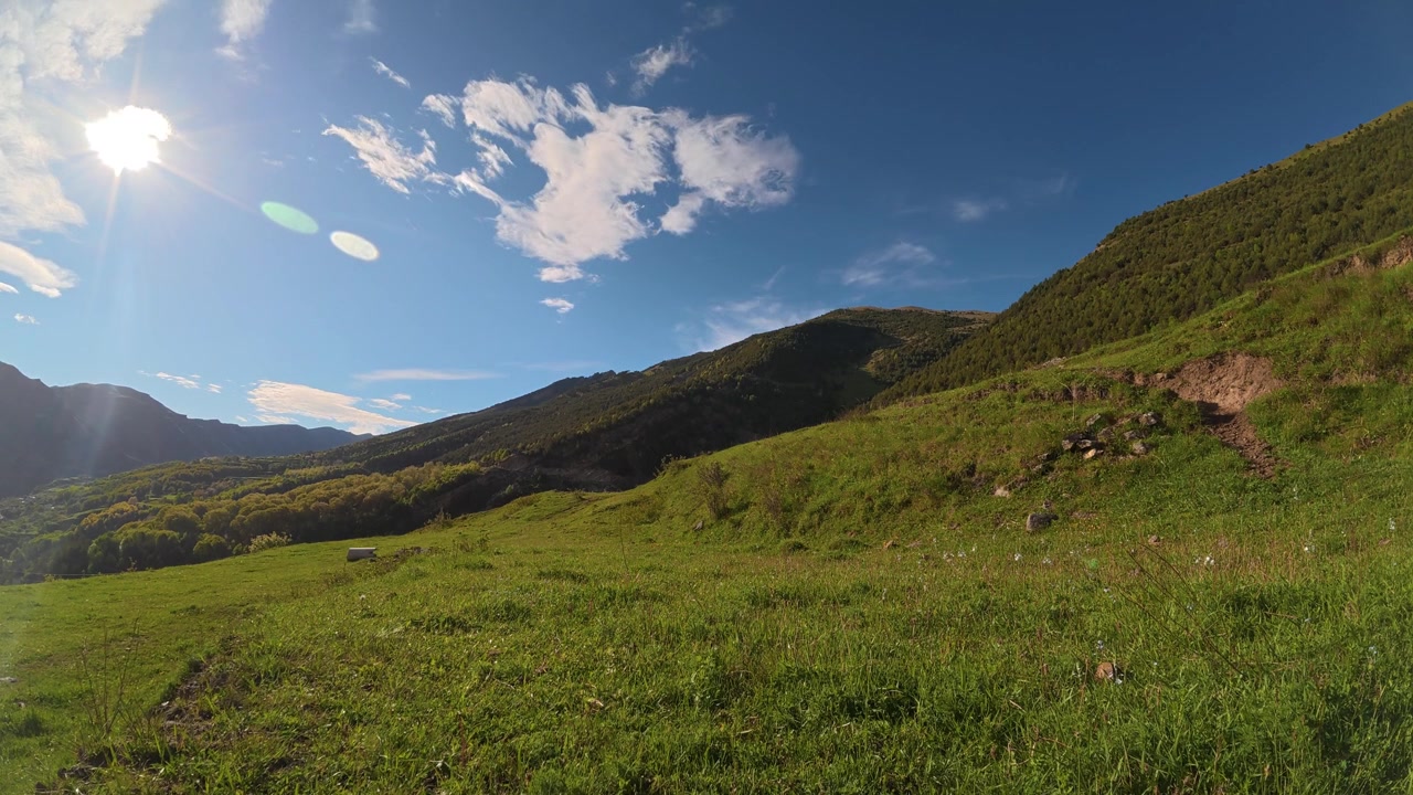 Un'ampia ripresa in time lapse cattura un vibrante paesaggio montano verde sotto un cielo azzurro brillante con nuvole che passano, con un bagliore solare sulla sinistra.