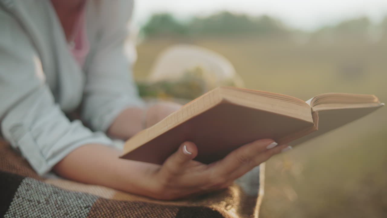 Primo piano della mano di una donna con un bracciale che gira una nuova pagina su una coperta all'aperto, illuminata da un bagliore dorato, con una vista sfocata di un cappello nelle vicinanze.