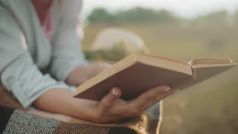 close-up of a woman hand with a bangle flipping to a new page on a blanket outdoors, highlighted by a golden glow, with a blurry view of a hat nearby