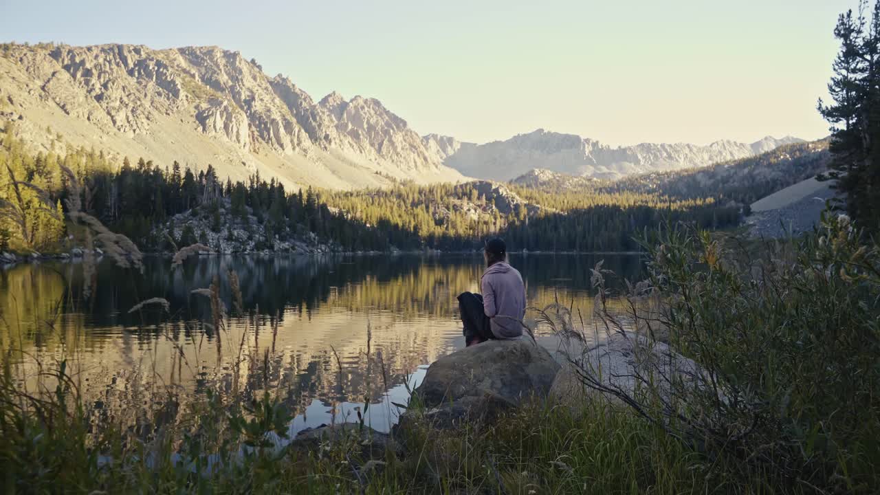Man sits by a serene mountain lake reflecting rugged peaks at sunset
