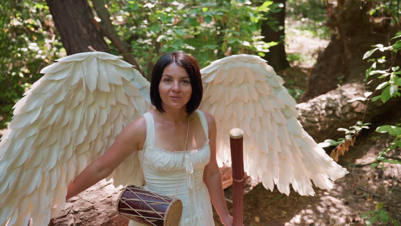 Woman dressed in white with large feathered wings standing in sunlit forest, serene expression glowing in natural light, embodying angelic presence and mystical calm among trees and soft foliage