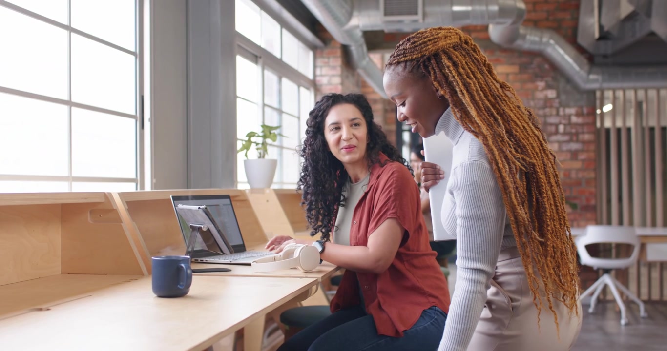 Diverse female coworkers at counter, braided woman carrying blue mug, leaning, reviewing tablet