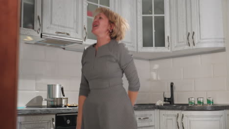 elegant lady in patterned dress jumps joyfully, dances with energy, pointing both hands sideways while grooving to music in kitchen, enjoying rhythm with passion, movement, and lighthearted spirit
