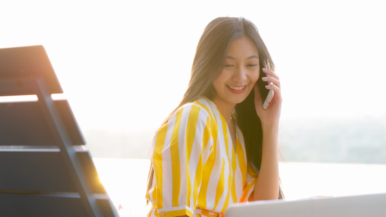 여름에 노란색 드레스를 입은 아름다운 아시아 여성이 수영장 근처에 앉아 전화로 이야기하고 노트북을 사용합니다. 행복한 여성 고객은 온라인 웹사이트 쇼핑 주문 배달을 확인하는 모바일 전화를합니다.