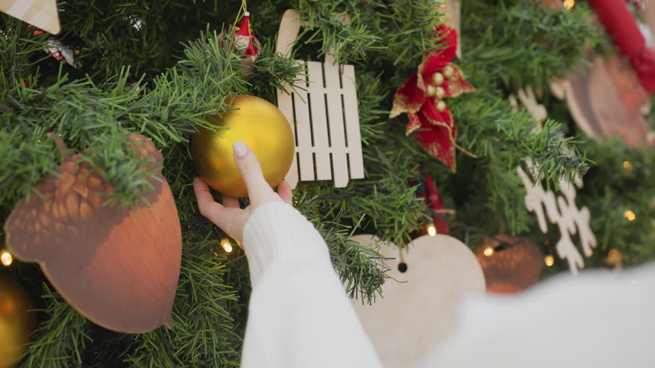 Close-up of woman in long sleeve delicately holding a golden Christmas ball from a decorated tree, showcasing a festive mood with a softly blurred background in a shopping mall