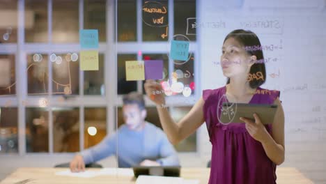 Mujer-De-Negocios-Asiática-Casual-Usando-Una-Lluvia-De-Ideas-En-Una-Tableta,-Tomando-Notas-En-Una-Pared-De-Vidrio-Por-La-Noche-En-La-Oficina