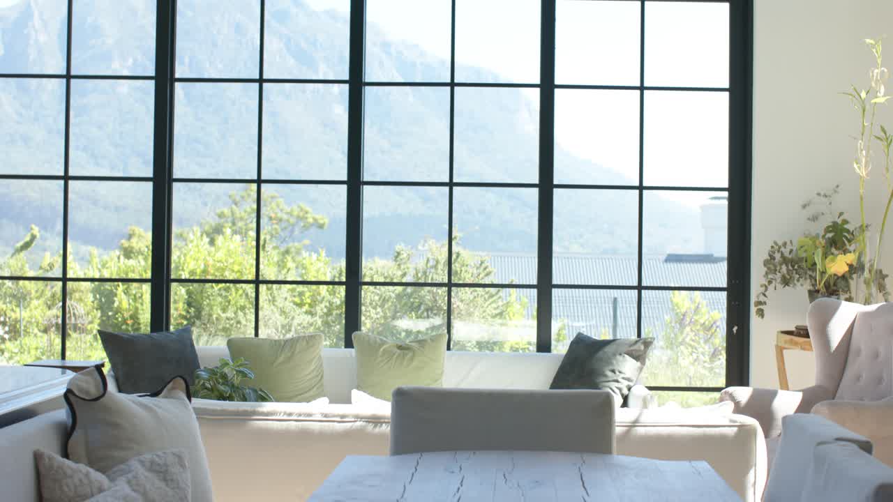 La cámara se inclina hacia arriba y se aleja para revelar una mesa de comedor rústica, un sofá y una vista de ventana soleada