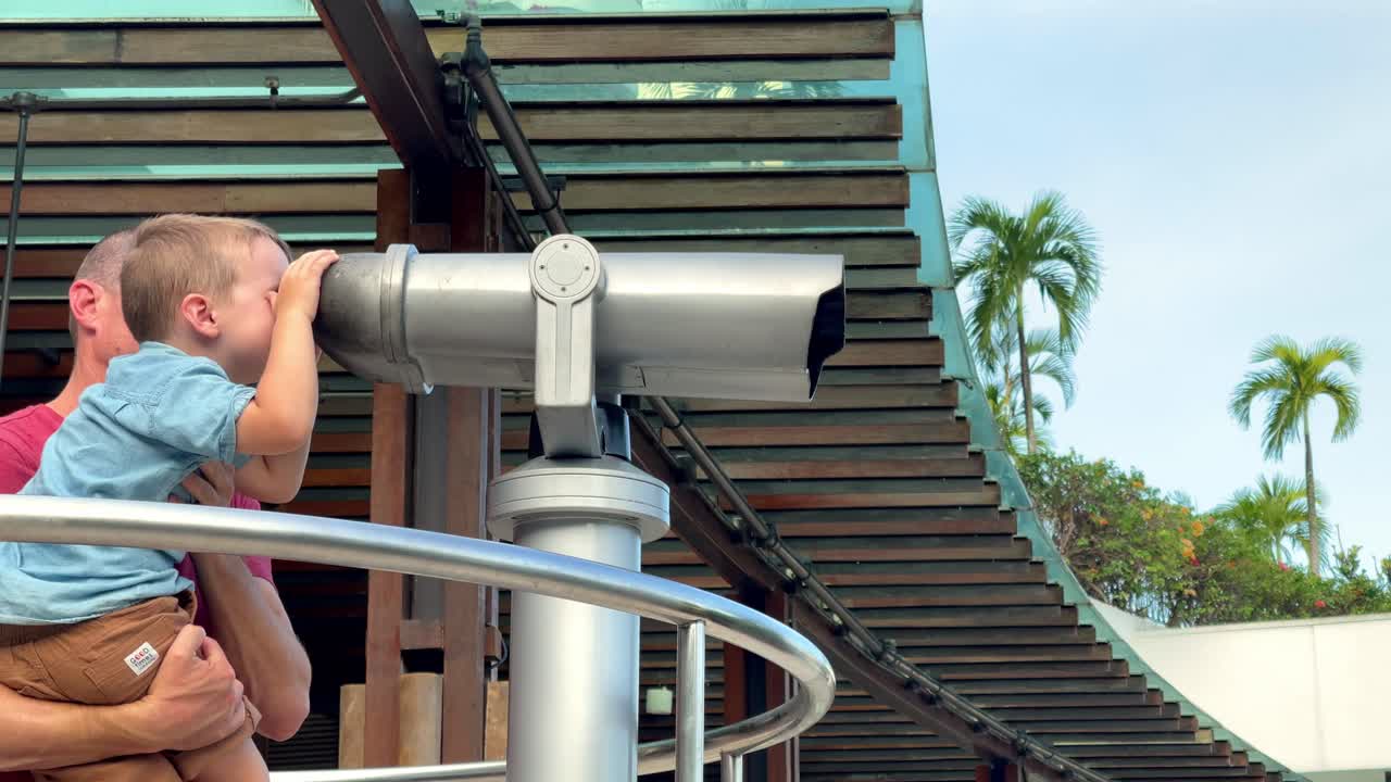 Man helps his young son to reach and use tower viewer binoculars at a tourist lookout, clip 2