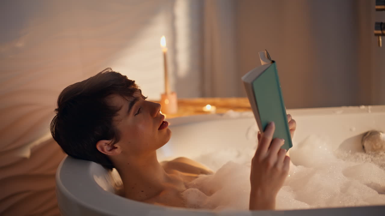 Bathing woman reading book in home bathroom closeup. Lady lying in candles light
