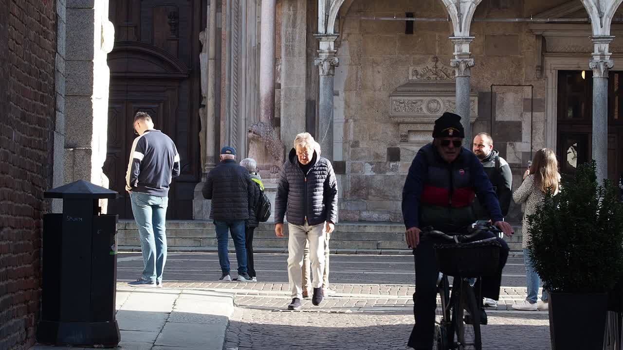 Elderly Man Rides Bicycle Past People Walking on Cobblestone Street in Historic European City