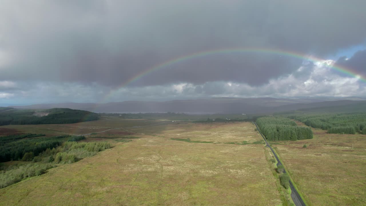 altas imágenes de drones aéreos bajo la lluvia rodando justo encima de una carretera larga y recta mirando hacia un arco iris brillante con nubes grises oscuras en el fondo, con campos, árboles y páramos en escocia