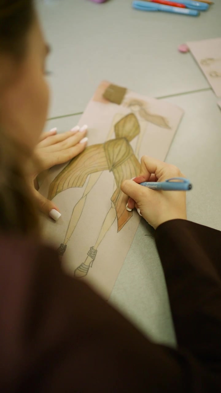 Primo piano di una stilista che disegna l'illustrazione di un abito strutturato dorato con matite colorate su un tavolo da lavoro in studio.