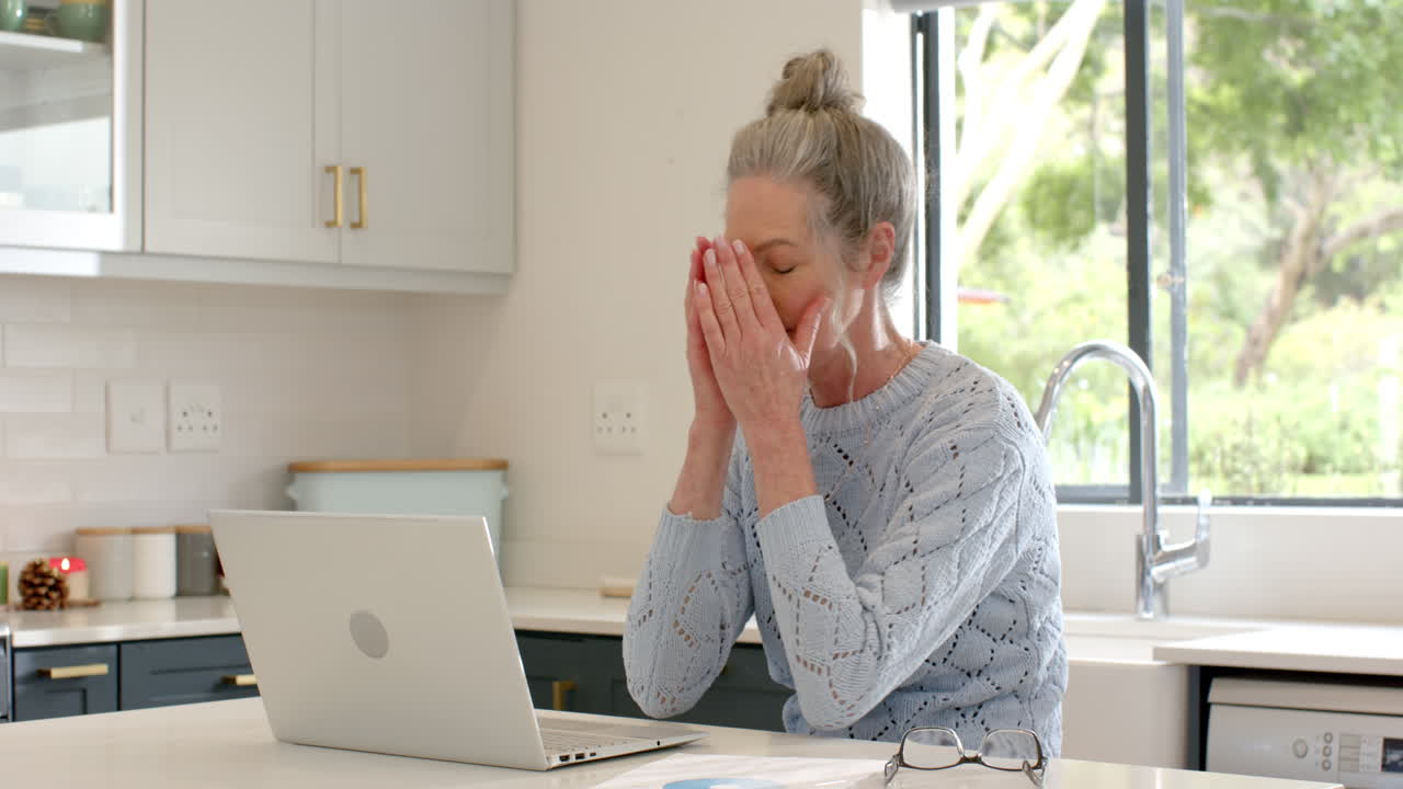 Mature woman feeling stressed at home using laptop in kitchen