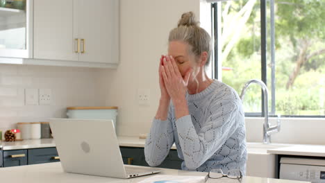 mature woman feeling stressed at home using laptop in kitchen