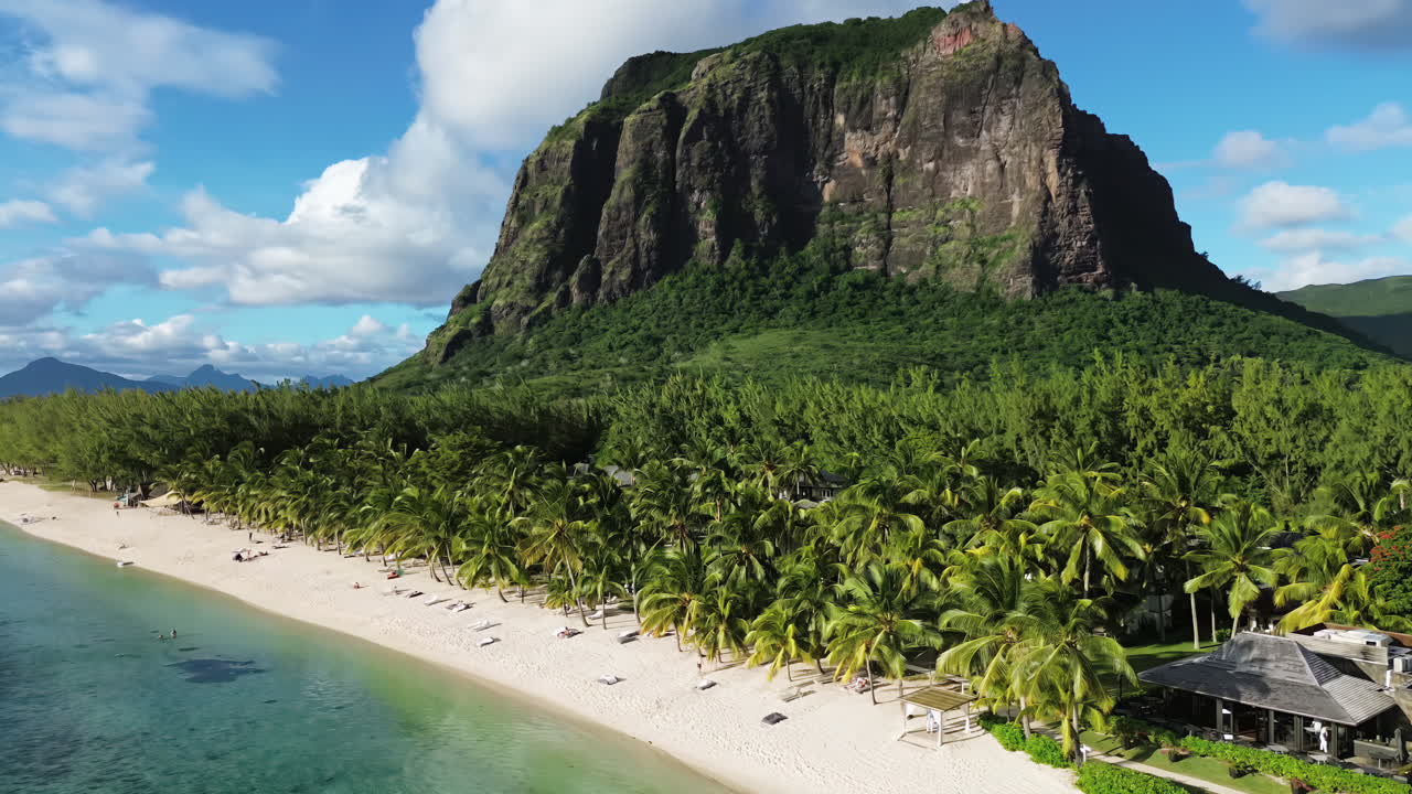 Vista aérea de drone ao longo de uma praia de areia branca ladeada por palmeiras na base da montanha Le Morne Brabant nas Maurícias, com água turquesa da lagoa e um resort tropical ao longo da orla.