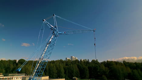 full view of a blue luffing construction crane against a blue sky with forest in background