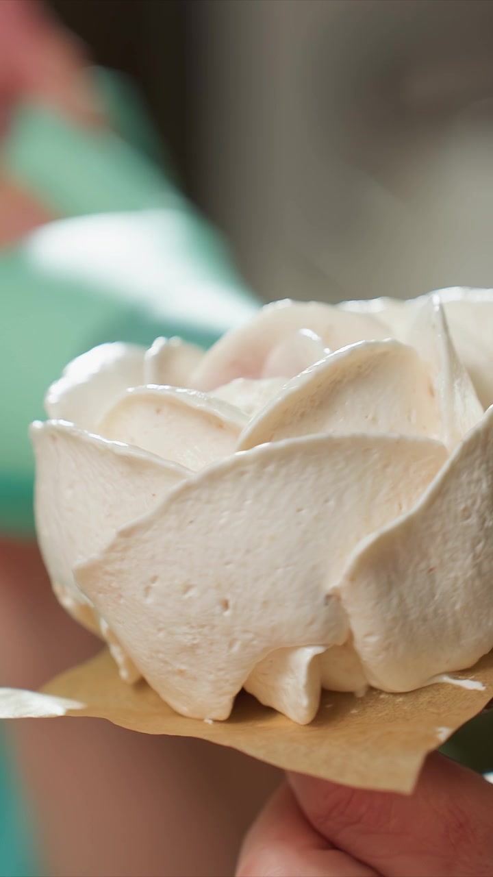 Dekat tangan memegang meringue putih berbentuk mawar. Tekstur lembut dan detail ditekankan saat objek diglasir. Pencahayaan lembut menyoroti permukaan halus sambil mempertahankan fokus