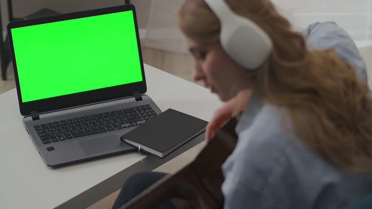 young woman is playing guitar sitting in front of laptop with green screen, conception of self-help guide or online rehearsing