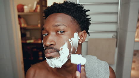 young black man looking into camera as shaving his face in bathroom