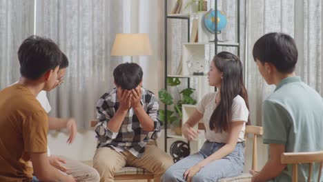 asian teen group sitting in chairs forming a circle discussing at home. teen students support a boy in plaid shirt not to crying