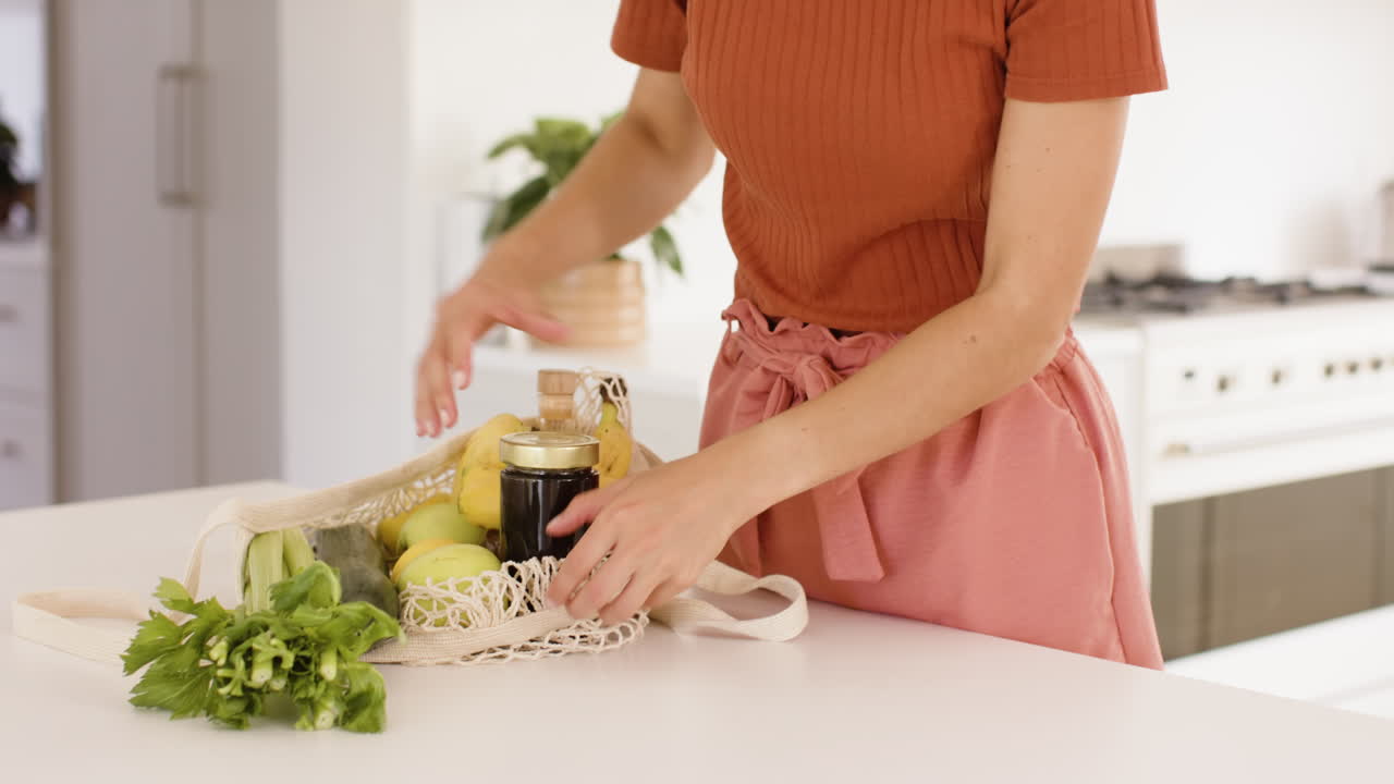 Woman unpacking groceries in kitchen, placing fresh produce on white countertop