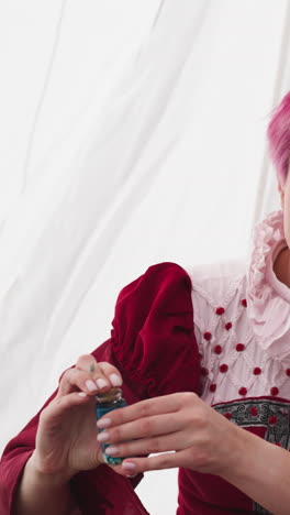 young witch corks small glass bottle with blue potion sitting at table. woman with short pink hair communicates pouring miraculous drink into bottles closeup