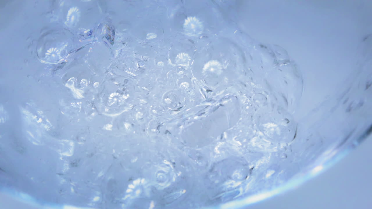 슬로우 모션 - 밝은 파란색과 흰색 배경이 있는 얼음 조각이 있는 유리잔에 t 물을 부었습니다.