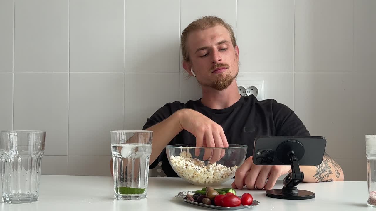 Молодой человек ест попкорн и смотрит смартфон дома