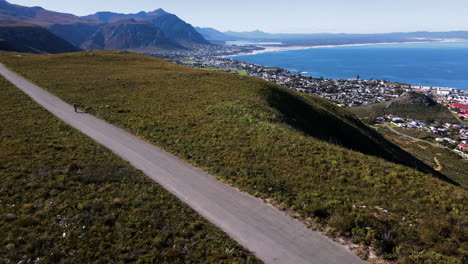 MTB-cyclist-on-scenic-mountain-road-overlooking-Hermanus-coastline,-aerial