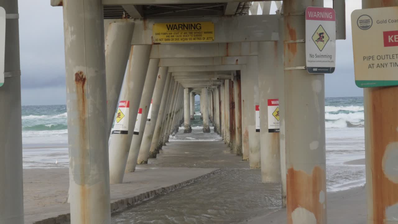 Scenic view under pier with warning signs: No Swimming, incoming waves during rainy day.