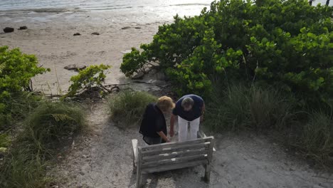 Ein-älteres-Paar-Setzt-Sich-Am-Strand-Auf-Eine-Bank,-Um-Sich-Auszuruhen-Und-Den-Sonnenuntergang-Zu-Beobachten