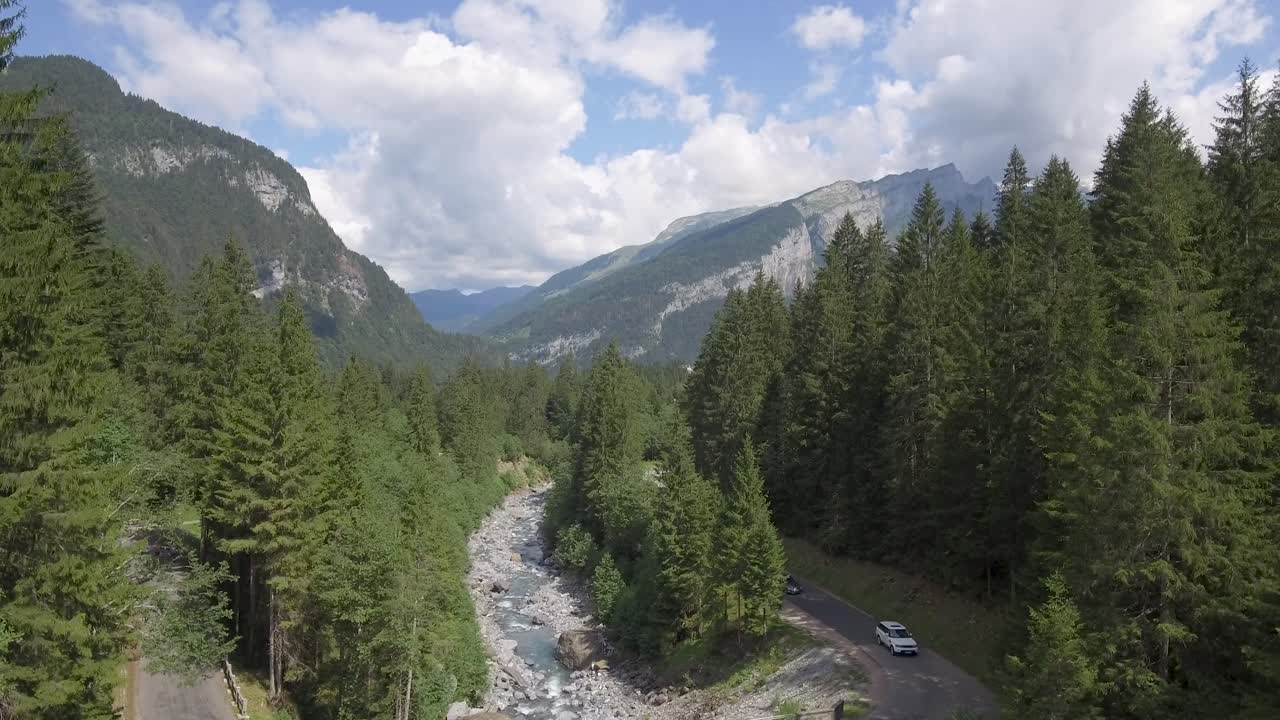Un drone vola sopra un ponte e una valle con un fiume di scioglimento glaciale, circondati da alte montagne e una lussureggiante foresta alpina verde
