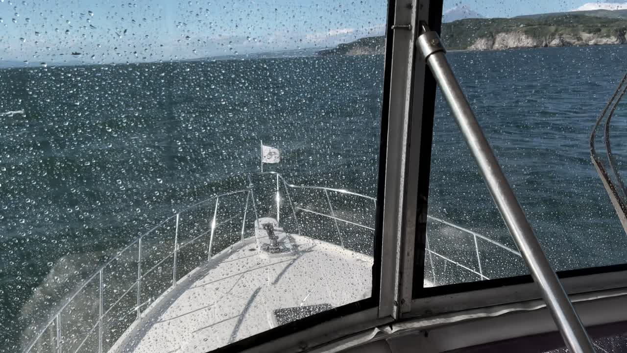 POV aus dem Inneren eines Bootes, das über den Bug durch ein regennasses Fenster blickt, während es an einem windigen Tag entlang der zerklüfteten Küste in der Region Kamtschatka, Russland, fährt