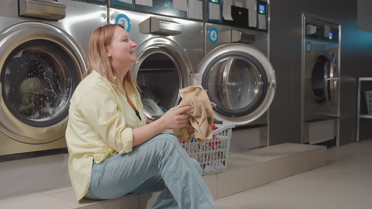 Mother playfully throws cloth to daughter, daughter tosses fabric into washing machine inside modern laundromat, washers with open door, basket of towels nearby, teamwork, hygiene routine