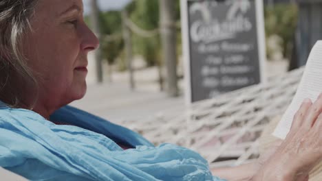 Happy-senior-caucasian-woman-sitting-in-hammock-reading-book-by-beach-bar,-in-slow-motion