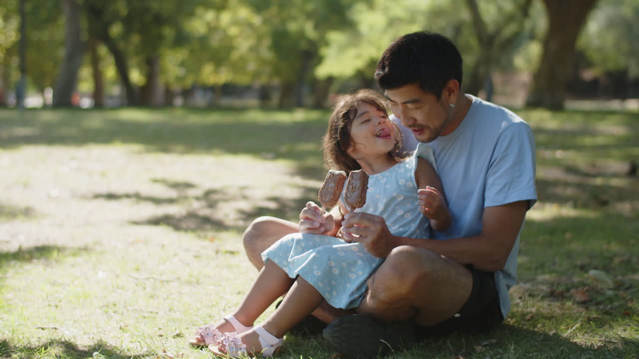 Cute little daughter sitting with father on grass and eating chocolate ice creams