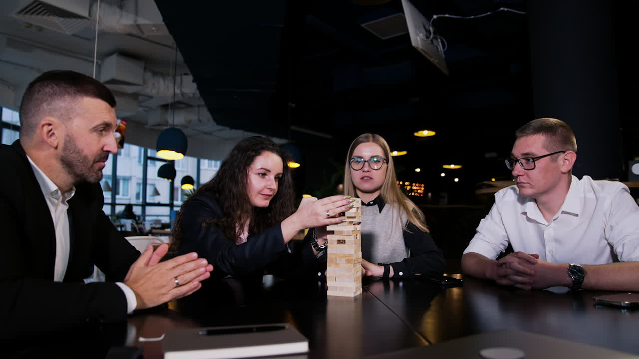 Four people sit at the table with jenga block pile on it. Dark-haired girl pulls the block very carefully while others watch her.